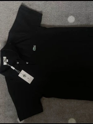 Svart pikétröja från Lacoste - Klassisk svart pikétröja från Lacoste med kort ärm och krage. Tröjan har knappar framtill och den ikoniska gröna krokodilloggan broderad på bröstet. Perfekt för en clean och sportig stil.