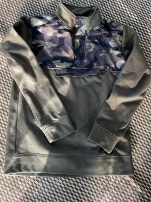Camo fleece-tröja från The North Face - Snygg fleece-tröja från The North Face med camo-mönstrat bröstparti och halv dragkedja. Tröjan är olivgrön med långa ärmar och hög krage. Perfekt för lager-på-lager och chill dagar. Broderad logga på bröstet.