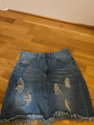 Blå jeanskjol med slitningar - Trendig blå jeanskjol med slitna detaljer och fransad nederkant. Kjolen har klassisk femficksdesign och bälteshällor. Perfekt för en avslappnad och cool stil.