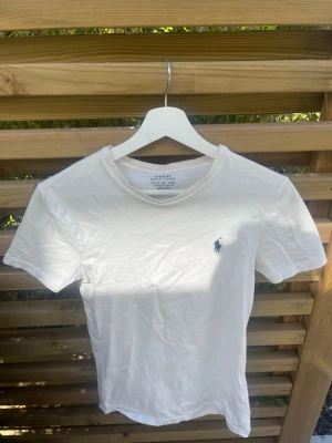 Vit t-shirt från Polo Ralph Lauren - Vit t-shirt från Polo Ralph Lauren med klassisk rund hals och diskret logga broderad på bröstet. Modellen är custom slim fit och tillverkad i mjuk bomull, perfekt för en clean och stilren look.