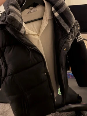  Burberry väst/jacka  - Säljer en svart pufferjacka från Burberry med klassiskt rutigt foder i huvan och dragkedja framtill. Jackan har en patch med texten 'Burberry London England' på ärmen och praktiska sidofickor. Perfekt för kalla vinterdagar och riktigt snygg streetstil.