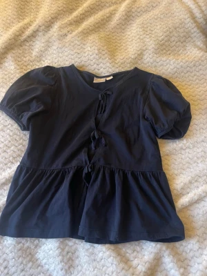 Mörkblå topp med knyt från Gina Tricot - Supersöt mörkblå topp från Gina Tricot med puffiga korta ärmar och knyt framtill. Toppen har en lätt utsvängd nederdel och är gjord i mjuk bomull. Perfekt för dig som gillar en feminin och trendig stil.