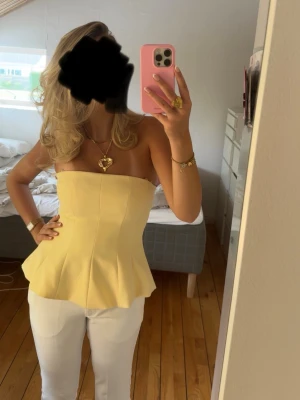 Gul peplum tubtopp - Snygg gul axelbandslös tubtopp med peplumskärning från Zara. Toppen har en figurnära passform upptill och utsvängd nertill. Det finns en liten fläck, se bild, men den märks inte om man inte vet att den finns.