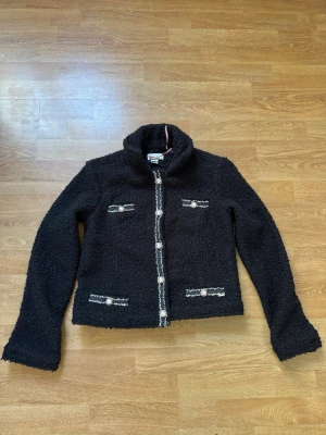 Holly & Whyte Lindex  - Svart teddyjacka från Zara med dekorativa knappar och detaljer i silver längs dragkedjan och fickorna. Jackan har hög krage och lång ärm, perfekt för att hålla värmen och ge en trendig look.