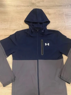Under Armour vindjacka - Snygg vindjacka från Under Armour i mörkblått och grått. Jackan har huva, hel dragkedja, bröstficka med dragkedja och logga på bröstet. Ärmsluten har tryckt text. Perfekt för blåsiga dagar och sportiga outfits.