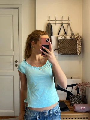 Ljusblå topp från Bisse Couture - Supersöt ljusblå topp från Bisse Couture med korta ärmar och vågig kant vid halsringningen. T-shirten har en feminin och enkel design som passar perfekt till jeans eller kjol. Mjuk och skön i bomullsmaterial.