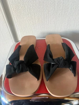 Svarta sandaler med rosett - Stilrena sandaler med breda svarta tygband och stora rosetter framtill. Sulan är i beige syntetmaterial med fyrkantig tå. Perfekta för en trendig och avslappnad look under varma dagar.