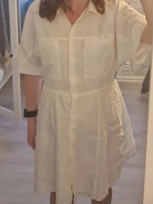 Vit skjortklänning i bomull - Säljer en vit skjortklänning med korta ärmar och två stora bröstfickor. Klänningen har knappar hela vägen framtill och materialet är i linne. Skriv vid intresse❤️