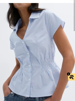 Blåvit randig blus med kort ärm - Snygg blus med blå och vita ränder, knappar framtill och markerad midja med smock. Klassisk krage och kortärmad modell i lätt bomullstyg. Perfekt till jeans för en fräsch look.