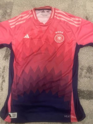 Tysklands rosa fotbollströja Adidas - Säljer en officiell Tyskland fotbollströja från Adidas i rosa och lila med zigzag-mönster. Tröjan har korta ärmar, rund hals och logga på bröstet. Tillverkad i lätt och ventilerande material med HEAT.RDY-teknologi. Helt ny! Pris kan diskuteras vid snabb affär!!