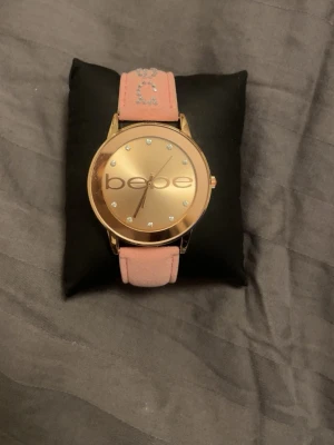 Guldig klocka från Bebe med rosa band - Stilren klocka från Bebe med stor guldig urtavla och diskreta stenar som timmarkeringar. Bandet är ljusrosa med glittriga stenar som bildar Bebe-loggan. Perfekt accessoar för att lyfta din outfit.