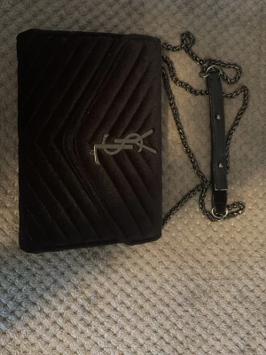 Svart sammet axelväska - Snygg svart axelväska i sammet med klassiskt silverfärgat logospänne framtill. Väskan har kedjerem med läderdetalj och quiltat mönster i V-form. Perfekt för dig som vill ha en lyxig och trendig accessoar.