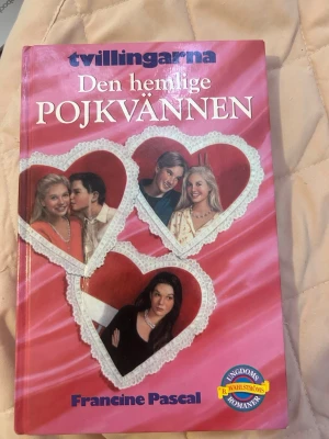 Tvillingarna: Den hemlige pojkvännen - En ungdomsroman av Francine Pascal ur serien Tvillingarna. Boken har ett rosa hårt omslag med hjärtformade bilder av karaktärer och vit spetsdekor. Perfekt för dig som gillar romantik och drama i skolmiljö.
