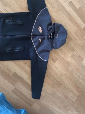 Svart Nike hoodie med dragkedja - Snygg svart hoodie från Nike med dragkedja framtill och två fickor med dragkedja. Hoodien har vita detaljer och Nike-logga på bröstet samt tryck på huvan. Tillverkad i mjukt material som passar perfekt till streetwear-stilen.