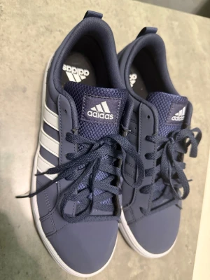 Blå Adidas sneakers med vita ränder - Snygga blå sneakers från Adidas med klassiska vita ränder på sidorna och vit sula. Ovandelen är i syntetmaterial och mesh, vilket ger både stil och komfort. Perfekta för dig som gillar en sportig och clean look.