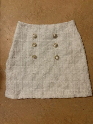 Vit kortkjol med knappar - Stilren vit kortkjol i quiltad struktur med sex dekorativa guldfärgade knappar framtill. Kjolen har en rak passform och känns både trendig och klassisk. Aldrig använd