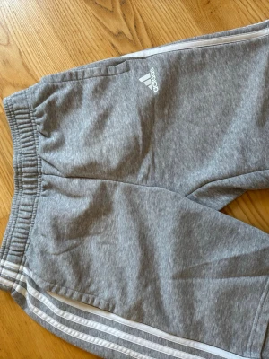Grå mjukisbyxor från Adidas - Snygga grå mjukisbyxor från Adidas med klassiska vita ränder längs sidorna och Adidas-logga på låret. Byxorna har resår i midjan och är gjorda i mjukt bomullsmaterial. Perfekta för chill eller träning.