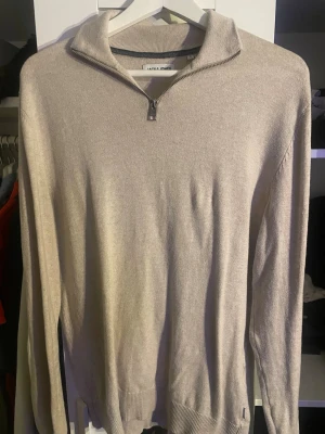 Beige halvzip-tröja från Jack & Jones - Snygg beige stickad tröja från Jack & Jones med halvzip och krage. Tröjan har långa ärmar och ribbade muddar vid ärmslut och nederkant. Perfekt för lager på lager och enkel att matcha med olika outfits.