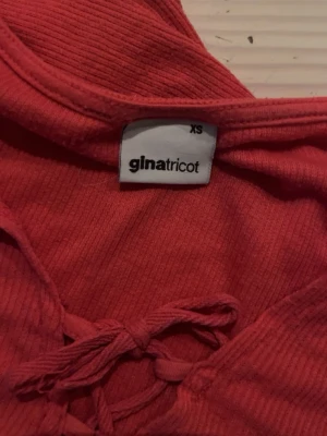 Röd ribbad topp från Gina Tricot - Säljer en röd ribbad topp från Gina Tricot i storlek XS. Toppen har korta ärmar och snygg snörning framtill vid halsen. Perfekt att styla med jeans eller kjol för en trendig look.