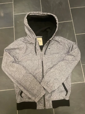 Grå vindjacka från Hollister - Säljer en gråmelerad vindjacka från Hollister med dragkedja framtill och svart innerfoder i huvan. Jackan har svarta detaljer vid fickorna och muddar vid ärmslut och nederkant. Perfekt för en avslappnad stil.