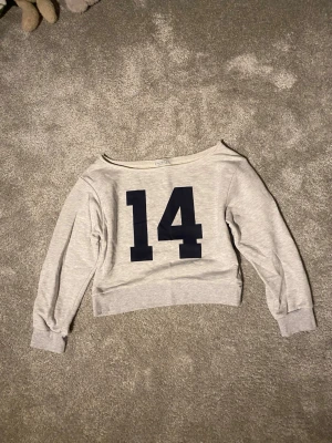 Grå croppad off shoulder hoodie Stradivarius - Snygg ljusgrå croppad hoodie från Stradivarius med stort mörkblått tryck av siffran 14 på framsidan. Tröjan har bred halsringning och långa ärmar. Perfekt för en off shoulder look