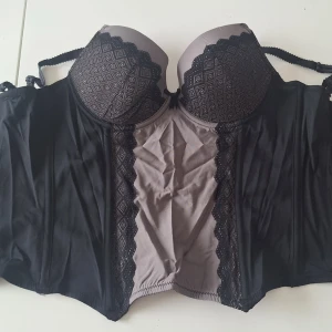 Vintage svart och grå spetskorsett - Snygg korsett i svart och grått med vadderade kupor och dekorativ spetsdetalj framtill. Axelbanden är justerbara och sidorna är i genomskinligt meshmaterial. Kuporna har ett diskret mönster och korsetten ger en feminin siluett. Många hyskor bak, därför passar den många. Använd 1 g. Köpt för 25 år sedan ca. Ber om ursäkt för att den är skrynklig, legat nerpackad.😊