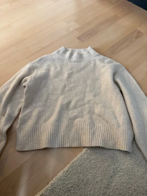 Beige stickad tröja Pull&Bear - Mysig beige stickad tröja från Pull&Bear med ribbad hög hals och bred ribbad kant nedtill. Tröjan har en avslappnad passform och långa ärmar, perfekt för lager på lager under kyliga dagar.