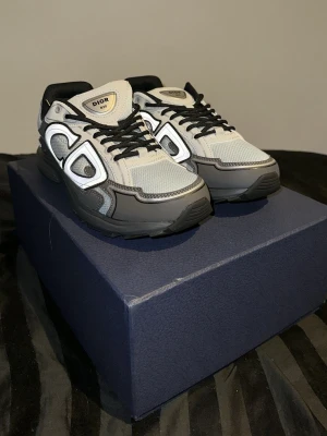 Dior B30 sneakers i grå och svart - Säljer ett par Dior B30 sneakers i grått och svart med vita detaljer har anvädvänt de 3-4 gånger. Skorna har en chunky sula, meshpaneler och reflekterande detaljer på sidorna. Klassisk Dior-logga på plösen och innersulan. Snygg och modern design som sticker ut.