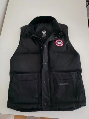 Svart dunväst från Canada Goose - Säljer en svart dunväst från Canada Goose med hög krage, två stora fickor framtill och klassisk logotyp på bröstet. Västen har en stilren design med knappar och dragkedja. Perfekt för kalla dagar och snygg att matcha med hoodie eller t-shirt.