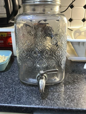 Glasbehållare med tappkran och lock - Stilren glasbehållare med vackert mönster, perfekt för drycker. Behållaren har en silvrig metalltappkran och ett matchande lock i rostfritt stål. Fyrkantig form och genomskinligt glas gör den enkel att fylla och servera ur. Perfekt till fest eller vardag i köket.