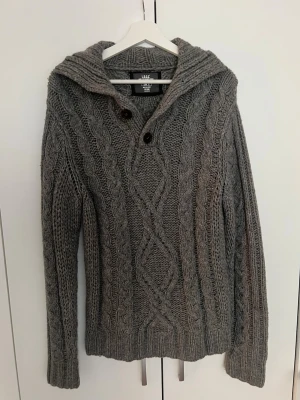 Grå kabelstickad hoodie  - Säljer en grå kabelstickad kofta från med knappar vid kragen och snyggt mönster både fram och bak. Tröjan har långa ärmar, ribbade muddar och en stor krage. Perfekt för dig som gillar en avslappnad men ändå stilren look.