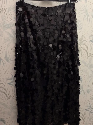 QNUS - Svart paljettkjol med stora paljetter, size S - Säljer en svart kjol täckt med stora, glansiga paljetter som ger ett riktigt statement-utseendeene. Kjolen har rak passform och är tillverkad i ett tyg som känns festligt och coolt. Perfekt för dig som vill sticka ut och älskar glitter och glam.                        Dragkedja bak.                                               Tags kvar 899kr