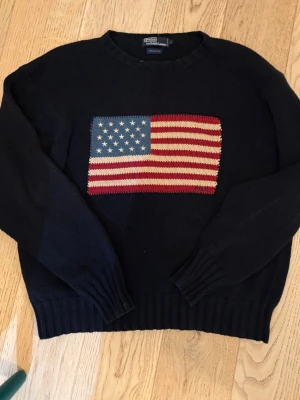 Svart stickad tröja Polo Ralph Lauren - Stickad svart tröja från Polo Ralph Lauren med amerikansk flagga broderad på bröstet. Tillverkad i 100% bomull och har ribbade muddar vid ärmslut och nederkant. Perfekt för dig som gillar klassisk och tidlös stil.