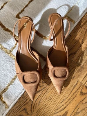 Bruna pumps med spetsig tå från Zara - Snygga bruna pumps från Zara i mockaimitation med spetsig tå och dekorativt spänne framtill. Skorna har smala remmar och en elegant siluett som ger en trendig look.