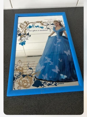 Disney spegel med blå ram och Cinderella - Säljer en dekorativ spegel med blå ram och motiv av Cinderella i blå klänning, omgiven av fjärilar och guldfärgade detaljer. Texten 'Once upon a moment...' finns på spegeln. Perfekt som väggdekoration för Disney-fans.