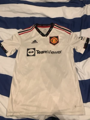 Manchester United bortatröja Antony - Snygg vit Manchester United bortatröja från Adidas med svarta och röda detaljer. Tröjan har klubbmärke, TeamViewer-logga och spelarnamn Antony med nummer 21 på ryggen. Tillverkad i lätt och ventilerande material, perfekt för fotbollsträning eller match.