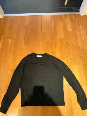 Svart sweatshirt från Calvin Klein Jeans - En stilren svart sweatshirt från Calvin Klein Jeans med rund halsringning och ribbade muddar. Tröjan har en diskret logga på ärmen och är tillverkad i mjukt bomullsmaterial. Perfekt för en clean och avslappnad look.