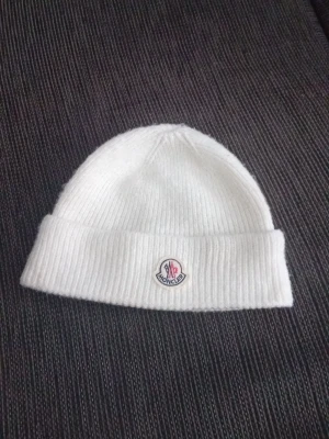 Vit mössa från Moncler - Säljer en stilren vit mössa från Moncler med uppvikt kant och klassisk Moncler-logga framtill. Mössan är gjord i mjukt material och passar perfekt för kalla dagar. Enkel att matcha med olika outfits.