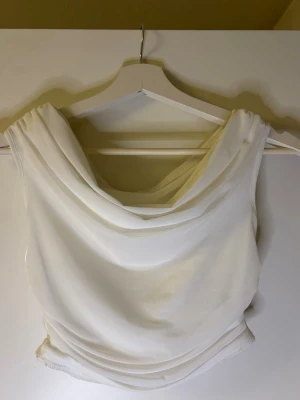 Vit ärmlös linnetopp med drapering - Säljer en stilren vit linnetopp med snygg draperad halsringning. Toppen är ärmlös och har en mjuk, följsam passform som ger en elegant look. Perfekt att matcha med jeans eller kjol för en trendig outfit.