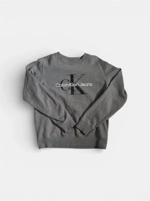 Grå sweatshirt från Calvin Klein Jeans - Säljer en grå sweatshirt från Calvin Klein Jeans med stort CK-logotryck på bröstet. Tröjan har rund hals, långa ärmar och ribbade muddar vid ärmslut och nederkant. Perfekt för en avslappnad och trendig look.
