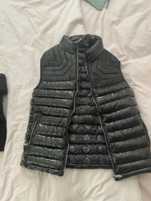 Svart dunväst från Philipp Plein - Snygg svart dunväst från Philipp Plein med glansig finish och coolt mönstrat innerfoder med döskallar. Västen har hög krage, dragkedja framtill och två fickor med dragkedja. Logga bak  och axelbandet. Perfekt för lager på lager. 