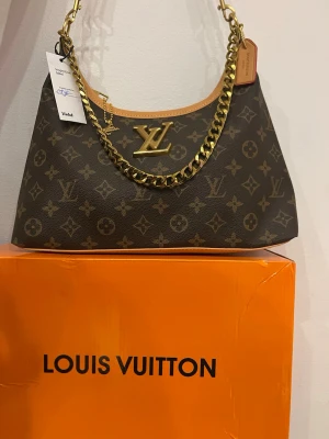 Louis Vuitton monogram axelremsväska - Snygg handväska från Louis Vuitton i klassiskt brunt monogrammönster. Väskan har en bred guldfärgad kedja som axelrem och stora LV-detaljer framtill. Tillverkad i canvas med läderdetaljer och har en exklusiv look som sticker ut.