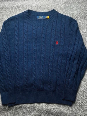 Polo Ralph Lauren - Kabelstickad svart tröja från Polo Ralph Lauren med rund halsringning och röd broderad logga på bröstet. Tröjan har ribbade muddar vid ärmslut och nederkant. Klassisk och tidlös design som passar till mycket.