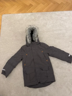 Mörkgrå jacka från polarn o pyret - Snygg mörkgrå jacka från Polarn O. Pyret med avtagbar huva och fuskpäls. Jackan har stora fickor framtill, reflexdetaljer på ärmar och rygg samt bruna detaljer vid fickorna. Perfekt för kalla dagar och riktigt skön att bära.