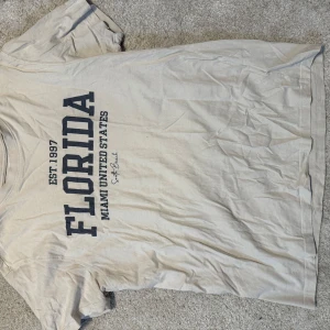 Beige Florida t-shirt från 157 - Säljer en beige t-shirt från 157 med trycket 'FLORIDA MIAMI UNITED STATES EST. 1997' i mörkblå text på framsidan. T-shirten är i 100% bomull och har en klassisk passform med korta ärmar. Perfekt för en avslappnad och cool stil.