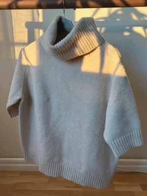 Beige stickad polotröja - Mysig ljusgrå stickad tröja med bred polokrage och halvlånga ärmar. Tröjan har ribbstickade detaljer vid krage, ärmslut och nederkant. Perfekt för lager på lager och en avslappnad stil. Materialet ser ut att vara mjukt och värmande. Man får med en matchade kjol!