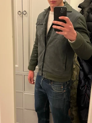 Mockajacka - Snygg mörkgrön jacka från Suede en Suède. Jackan är i faux suede material och ull blandning med ribbade muddar och krage. Fodrad på insidan och tål kyla bra. Jackan har två bröstfickor med lock och knappar samt två sidofickor. Stängs knappar framtill. Perfekt för dig som vill ha en stilren och modern look. Obs jackorna säljs helt nya och finns i storlekarna XS, S och M. Priset är fast. Hör av er vid frågor, mvh//David