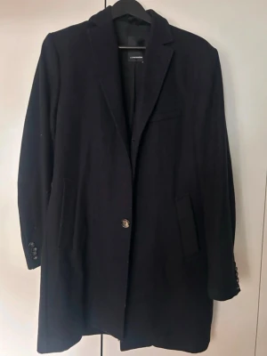 Svart ullkappa från J.Lindeberg - Stilren svart kappa från J.Lindeberg med klassisk krage, två fickor framtill. Kappan har en rak passform och är tillverkad i ullblandning, perfekt för kyliga dagar. En knapp saknas i mitten på kappan 