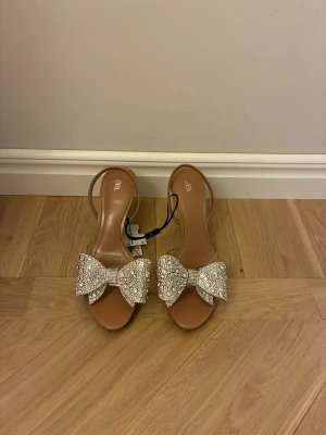Beige klackskor med strassrosett från Zara - Säljer ett par snygga klackar från Zara i beige med transparenta detaljer och en stor glittrig rosett framtill täckt av strass. Skorna har öppen tå, smal klack och slingback-rem. Perfekta för dig som vill sticka ut med lite extra bling. Nya, aldrig använda.