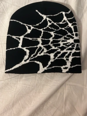 Svart mössa med spindelnätsmönster - Cool svart mössa med vitt spindelnätsmönster runt om. Perfekt accessoar för dig som gillar streetwear och vill sticka ut. Mössan är mjuk och stretchig, passar de flesta huvudstorlekar. Materialet känns som bomull eller akryl.